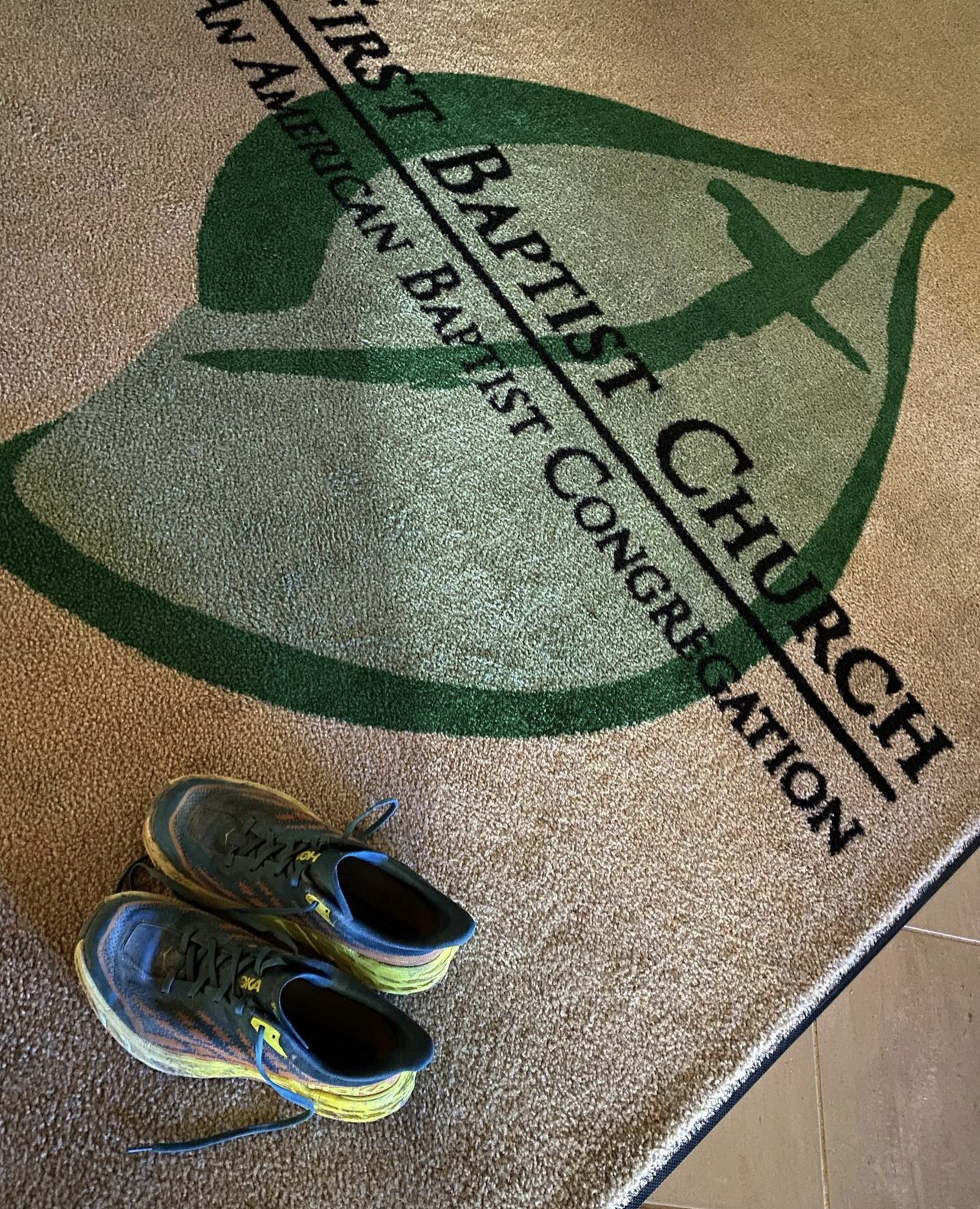 A photo of Pastor Matt's blue, orange, and yellow trail running shoes, on a rug with a leaf logo that reads "First Baptist Church, An American Baptist Congregation."